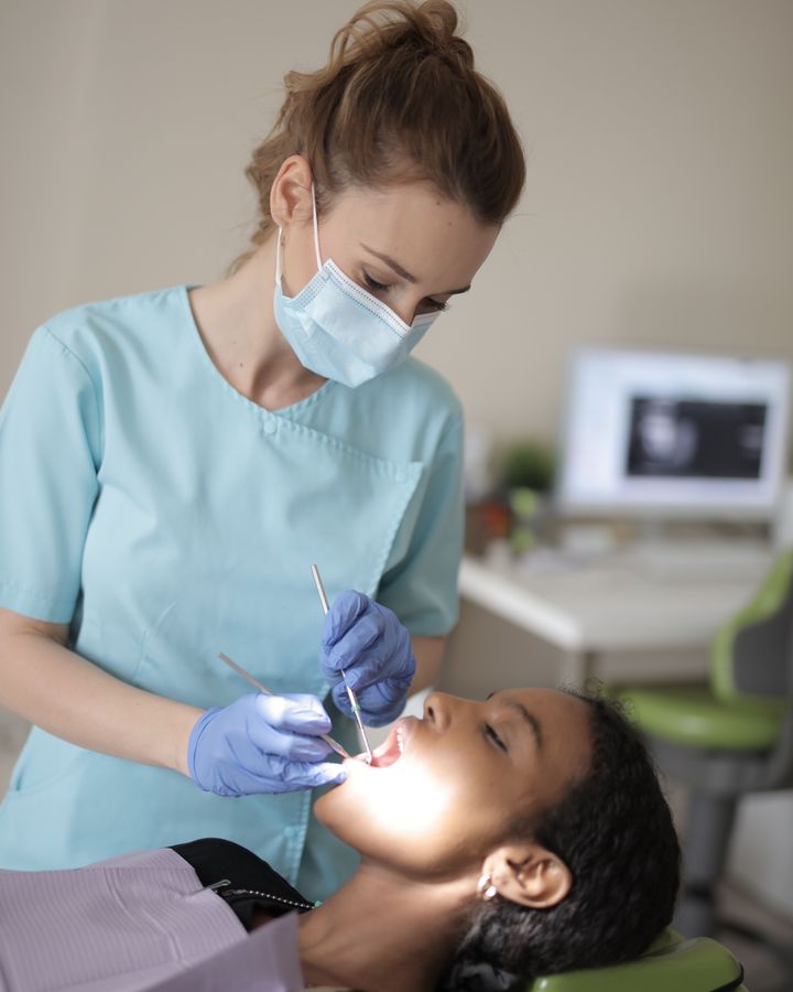 A dentist in teal scrubs examining a patient's teeth under a dental light, a digital scan visible on a monitor behind her