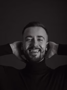 Black and white portrait of a bearded young man smiling, hands framing his face